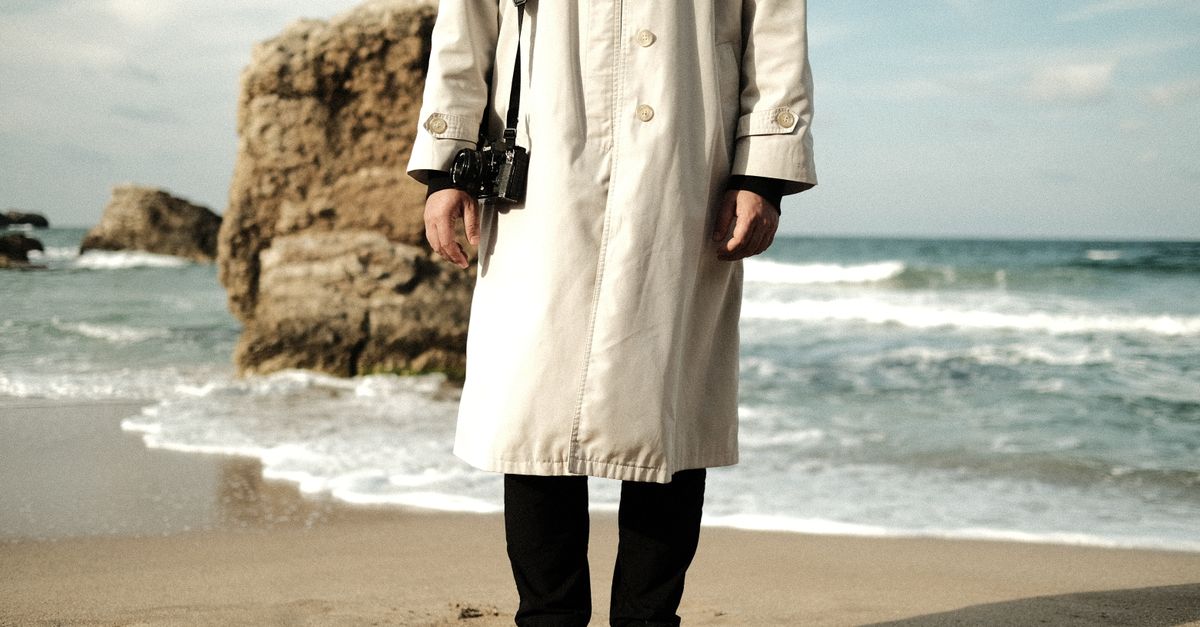 A stylish photographer in a trench coat stands at the seashore holding a camera.