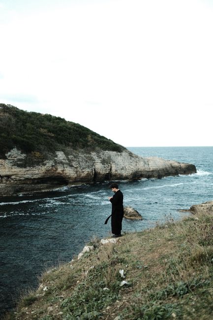 A person in a black coat stands alone on a rugged cliff overlooking the sea, with waves crashing below.