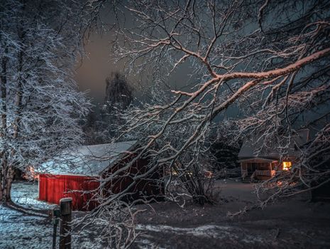 A red barn and house illuminated at night amidst snow-covered trees, creating a serene winter landscape.
