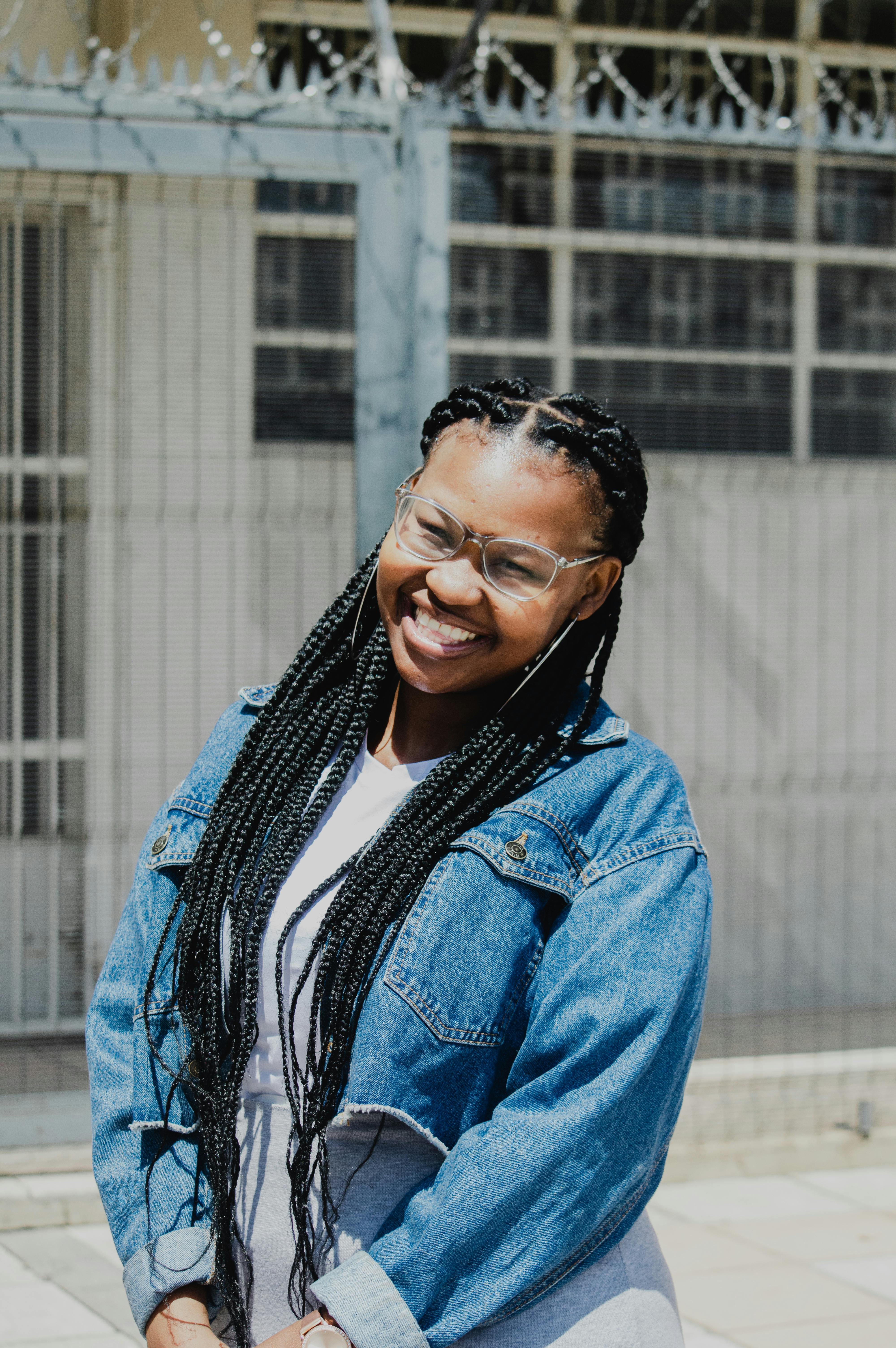 Portrait of a black woman with braids and eyeglasses, smiling brightly outdoors.