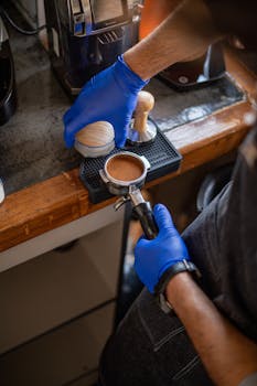 A barista wearing gloves preparing espresso using a coffee tamper and portafilter in a café setting.