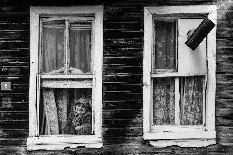 Elderly Woman Looking Out The Window In An Old Wooden House 