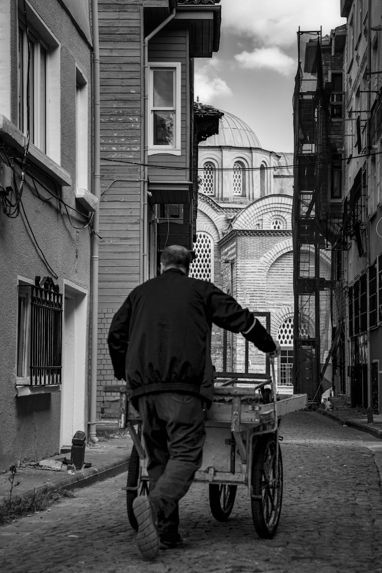 Back View Of A Man Pushing A Cart On A Cobblestone Street In City 