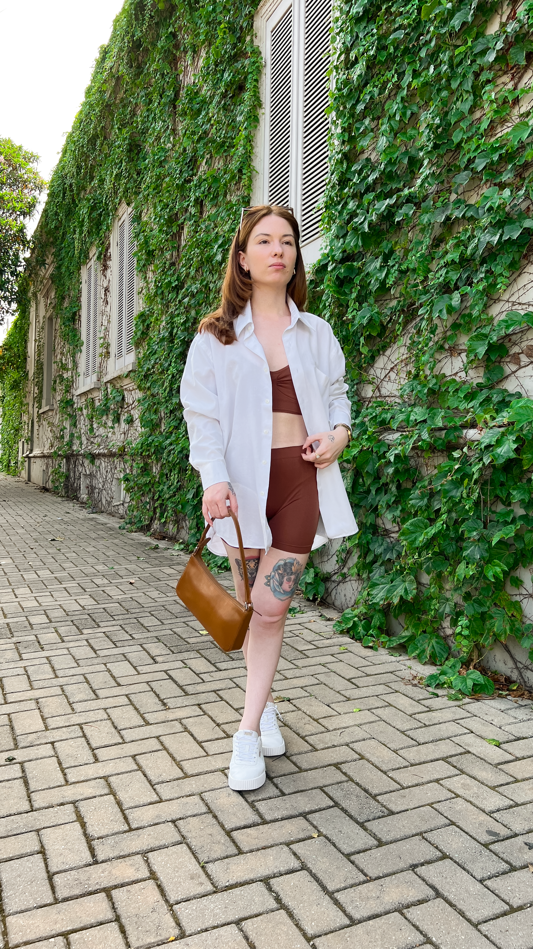 Mulher fashion em roupa casual caminhando ao lado de uma parede coberta de hera em um dia ensolarado.