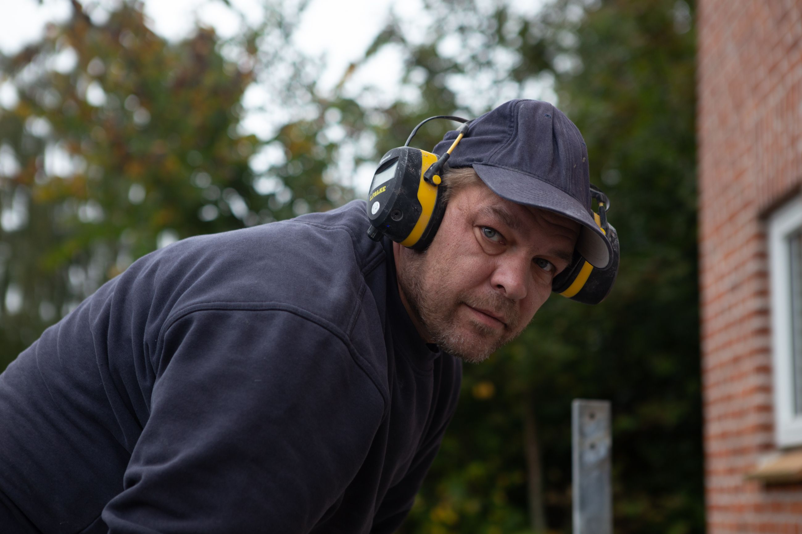 A man in protective headphones outdoors, leaning forward, wearing casual attire with a focused expression.