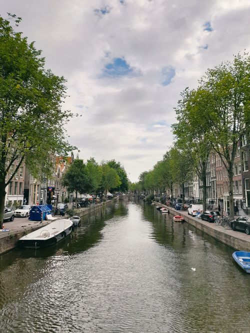 Free Scenic view of Amsterdam canal with boats and trees lining the banks under cloudy skies. Stock Photo