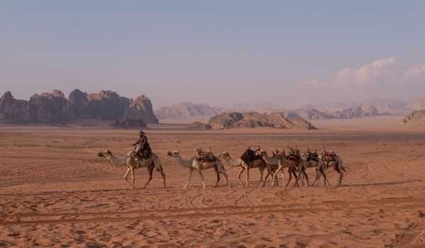 A camel caravan traverses the sandy desert near towering rocky formations, under a clear sky.