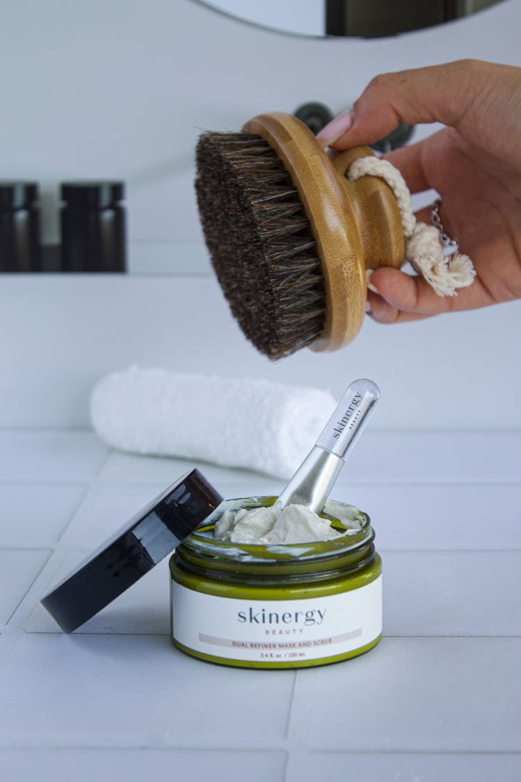 Close-up Of Woman Holding A Body Brush Above A Container With A Skincare Product