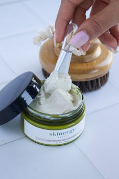 Close-up of a hand scooping a skincare product from an open jar, showing a brush in the background.