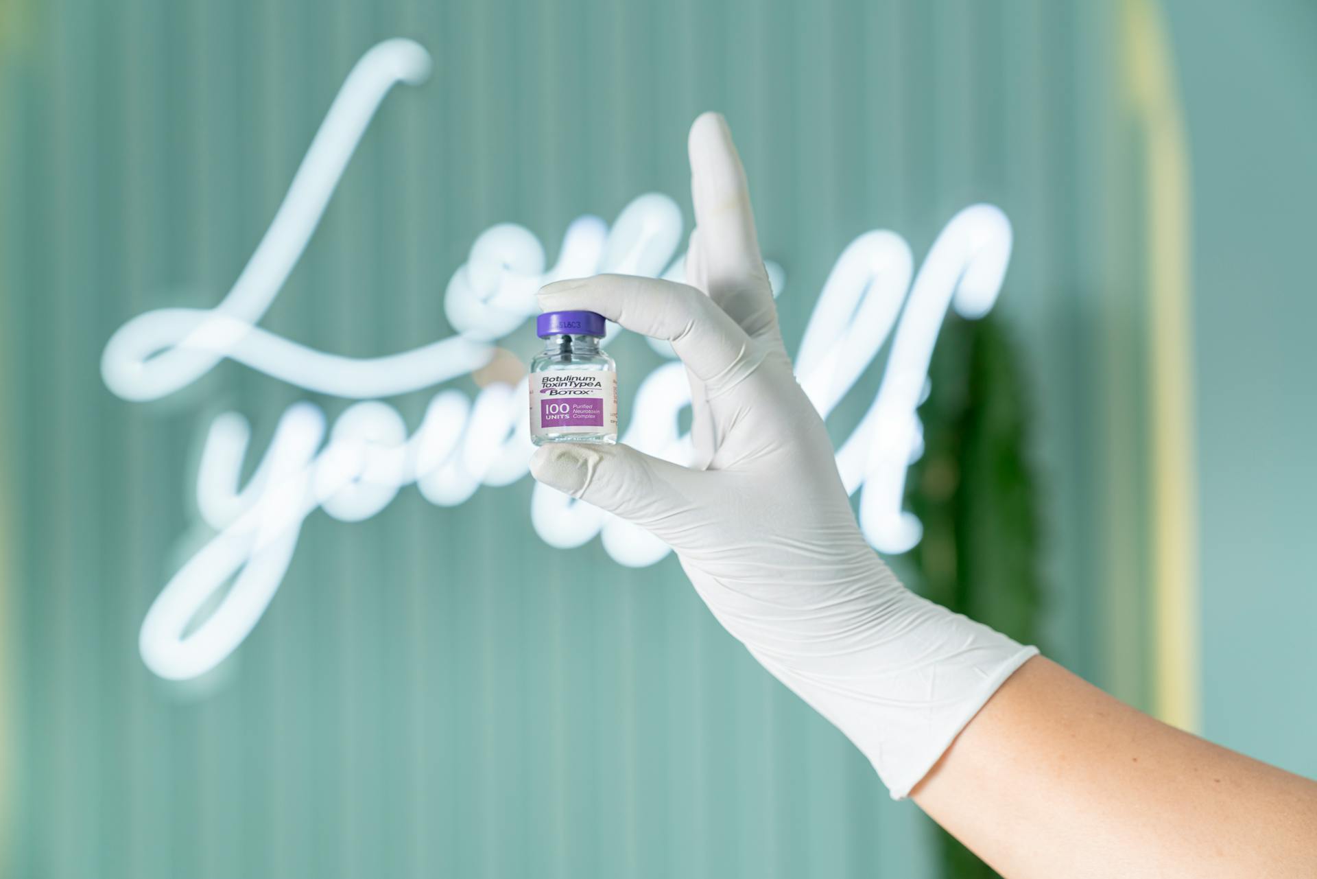 A gloved hand holds a Botox vial against a soft green background with neon lights.