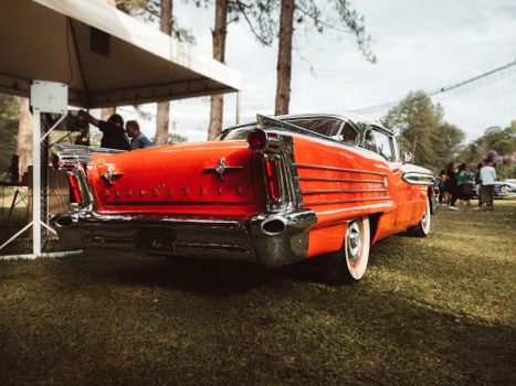 Rear view of a vintage orange car showcased outdoors at an auto show with people in the background.