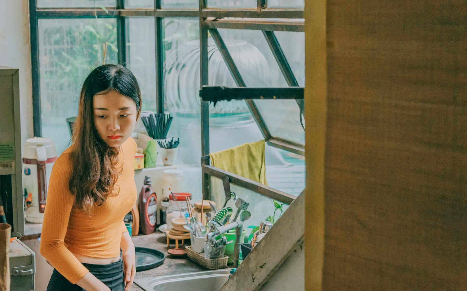 A young woman in a bright kitchen washes dishes by a sunlit window.