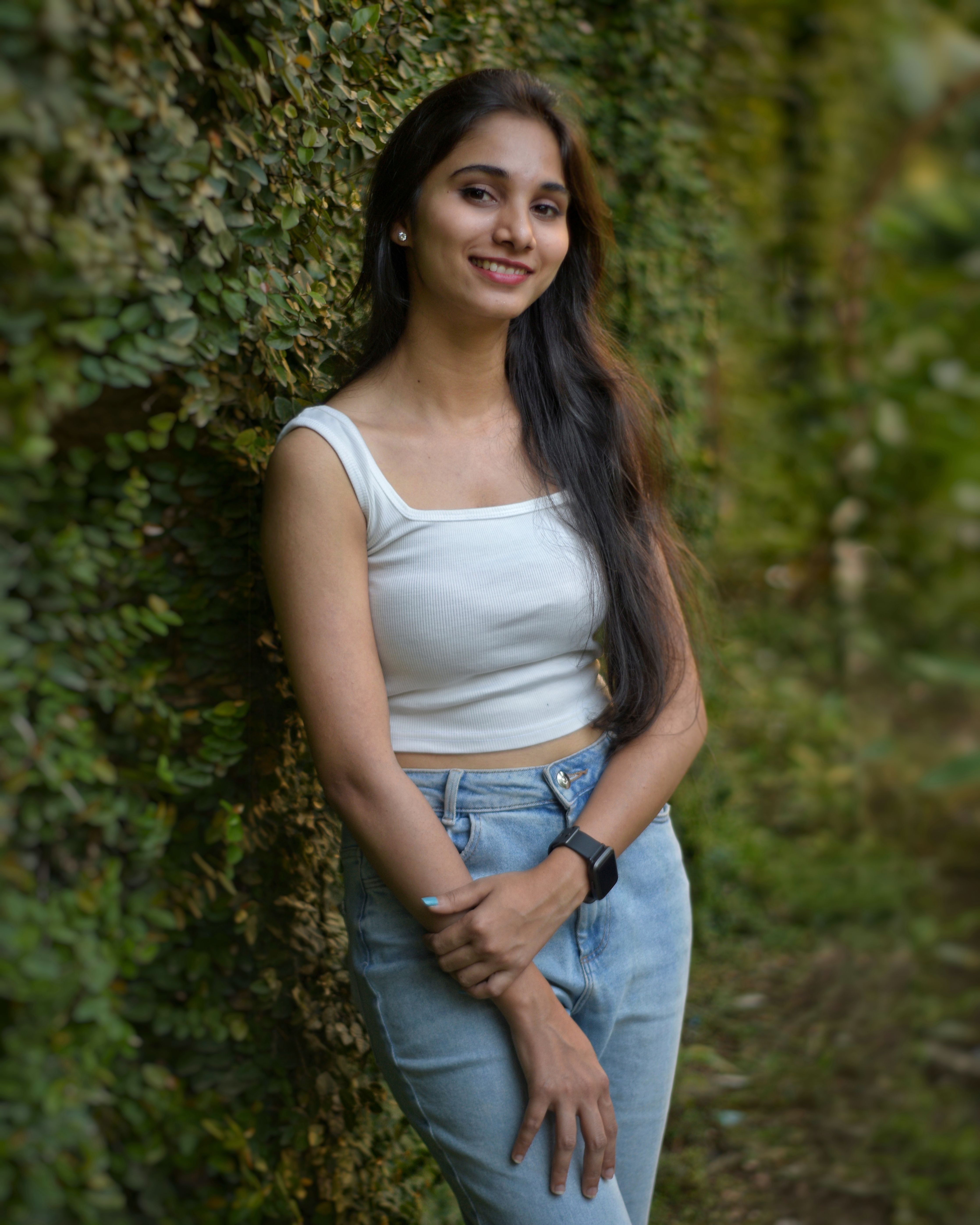 A young woman in a white tank top smiling, standing outdoors against a lush green background.