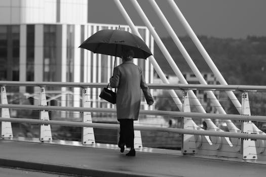 Black and white image of a person holding an umbrella walking across a bridge.
