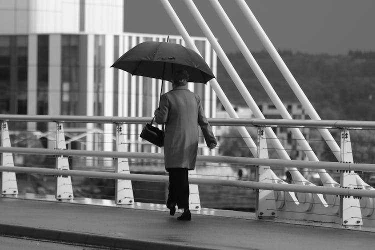 Grayscale Photo Of A Person Holding An Umbrella While Walking On The Bridge