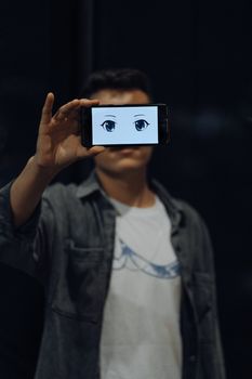 A person holds a smartphone displaying anime eyes, obscuring their face, wearing a denim jacket.. pexels, person, holds