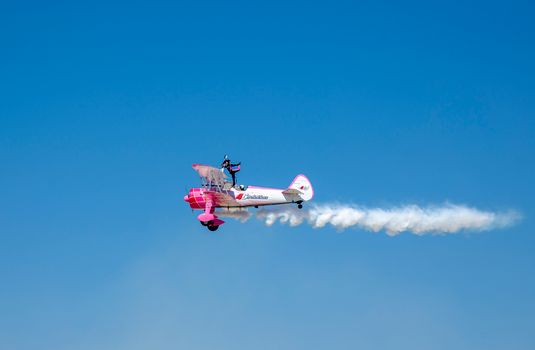 A stunt performer balances on a flying Boeing Stearman biplane against a clear blue sky, showcasing thrilling aerobatics.