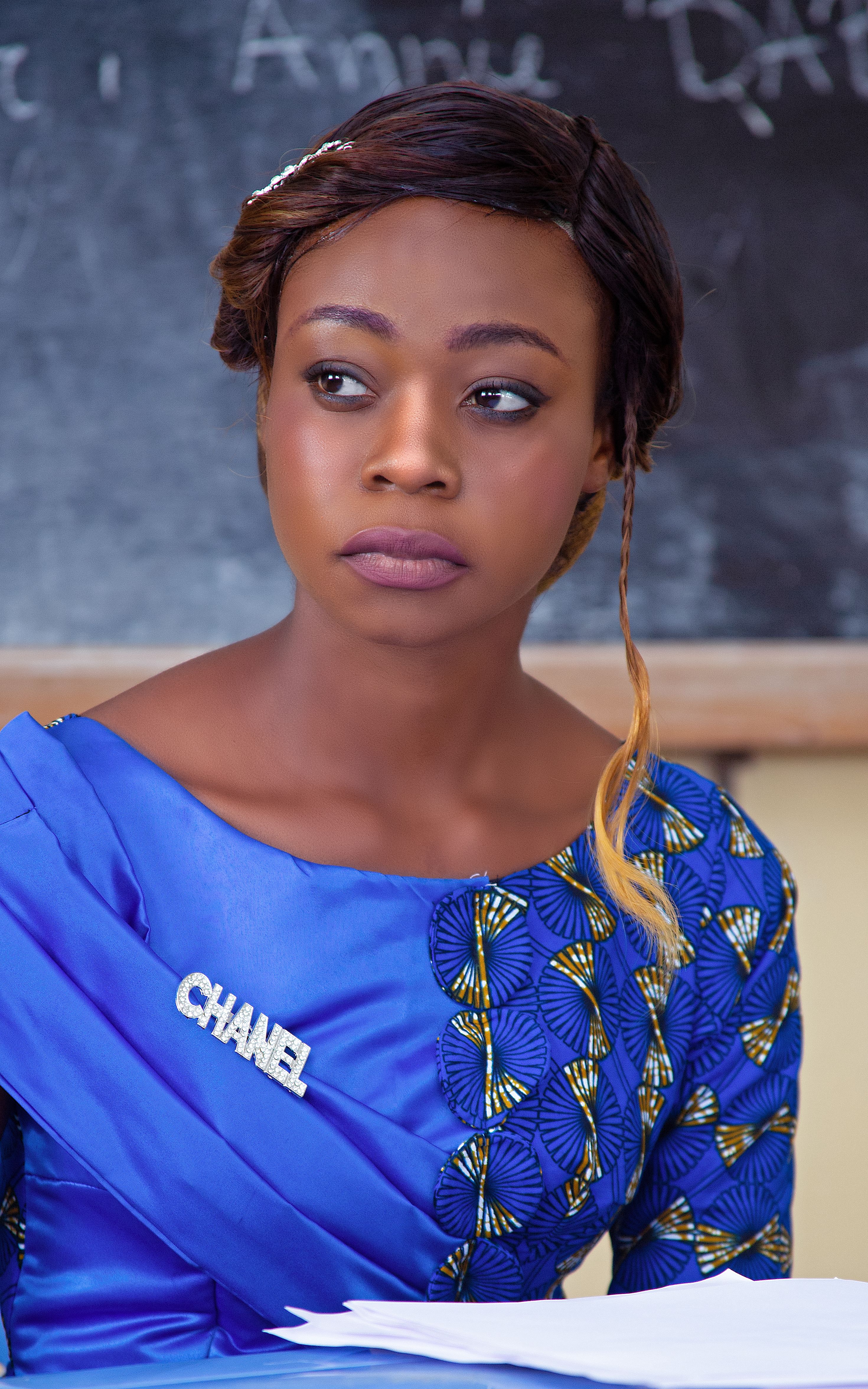 Stylish African woman in blue traditional outfit with Chanel brooch seated in front of a blackboard.