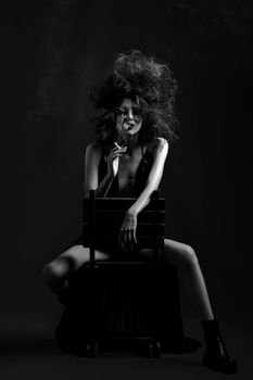 Dramatic black and white portrait of a woman sitting on a chair, smoking with hair styled wildly.