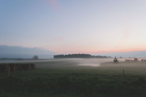 Free Serene misty field at dawn with hay bales and a tractor in soft light. Stock Photo