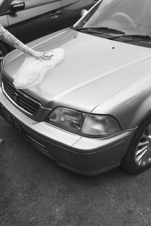 Grayscale Photo of a Person in Laced Long Sleeve Holding Veil on Car Hood