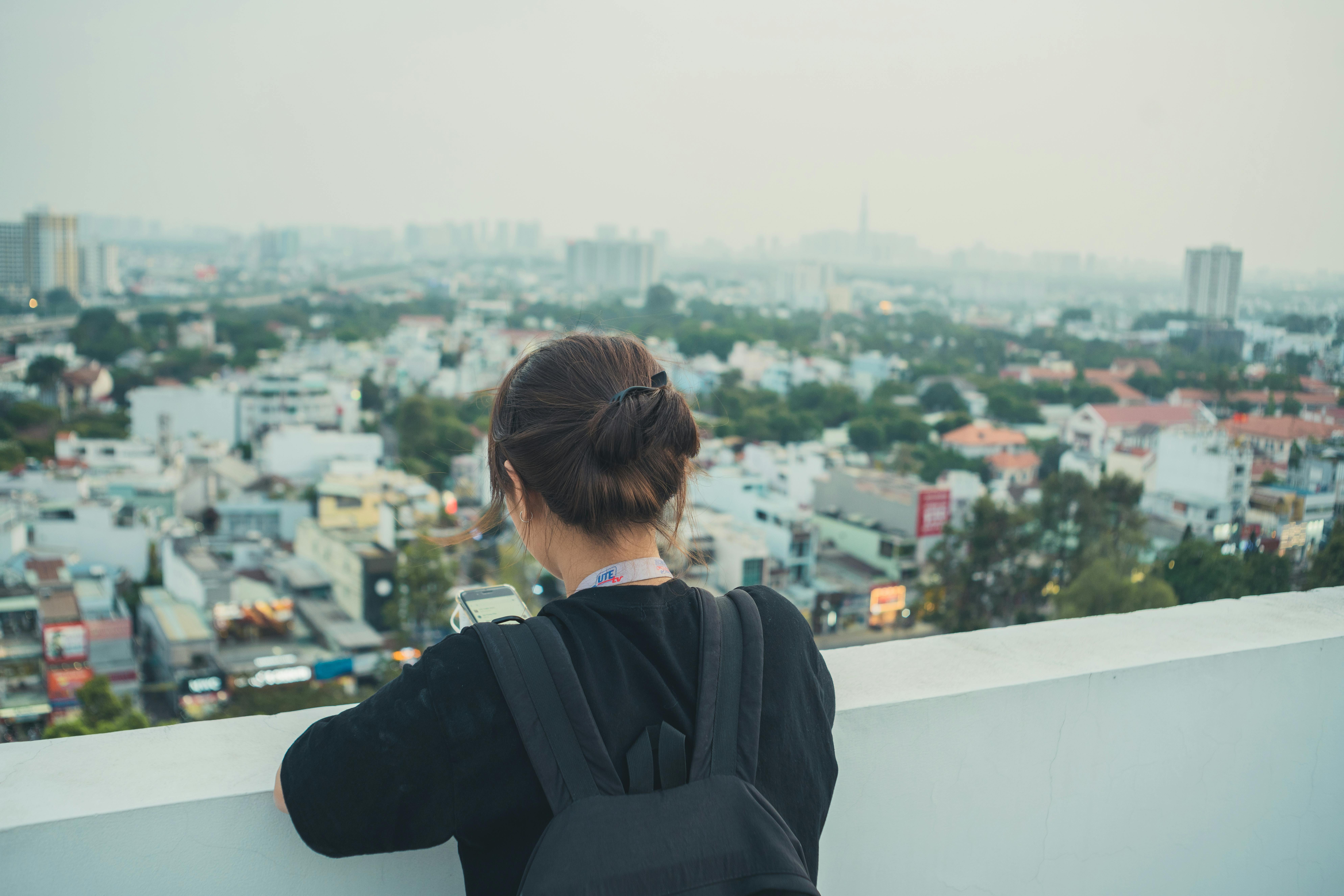 A woman with a backpack stands on a rooftop, using her phone to capture the urban cityscape view.