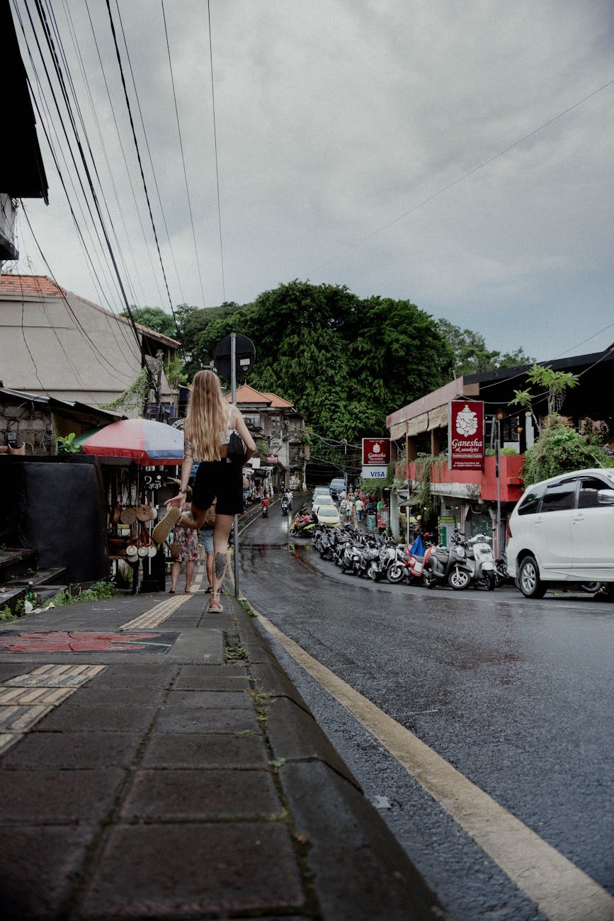 A woman walks along a wet street in a bustling urban area, showcasing daily life and vibrant shops.