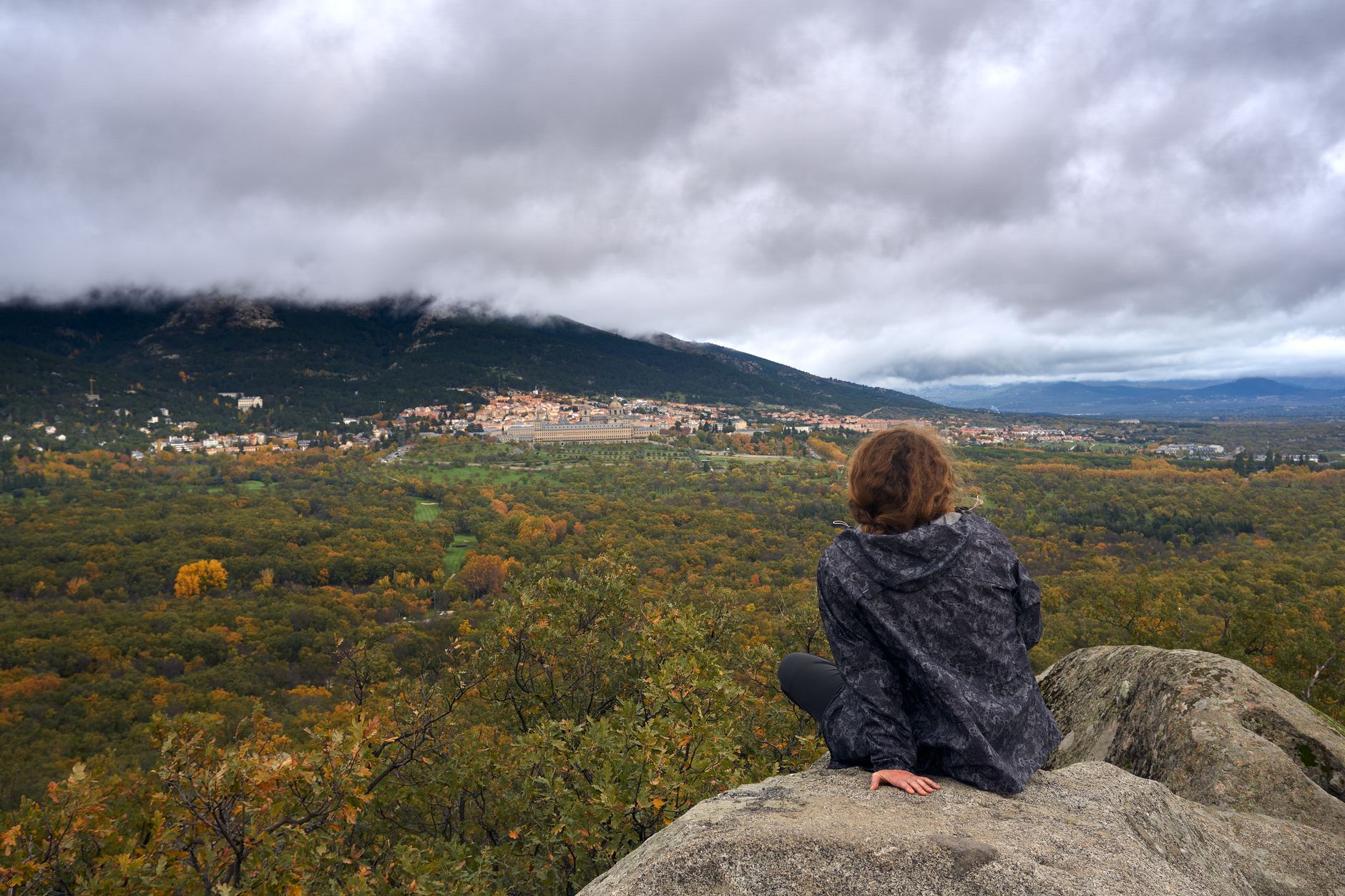 A person sits on a rock overlooking a vast forest with autumn colors under a cloudy sky.