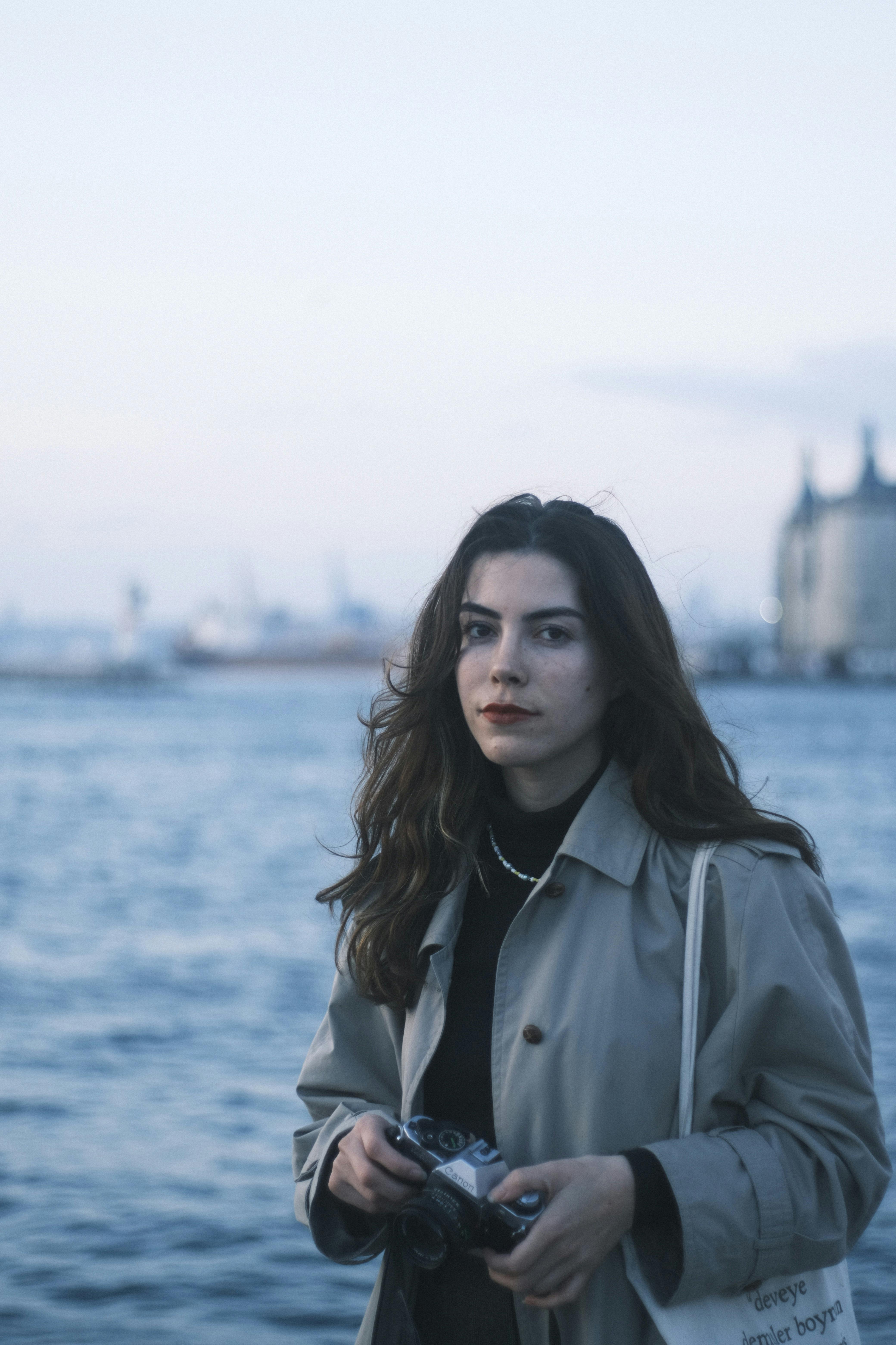 A pensive woman in a trench coat stands by the sea during twilight, holding a camera.