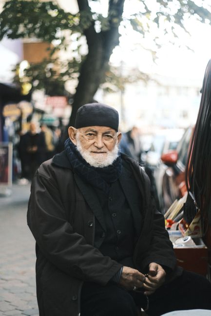 Portrait of an elderly man with a beard wearing a cap, sitting on a city street with a contemplative expression.