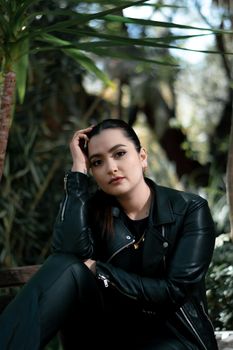 Portrait of a woman in a leather jacket sitting outdoors amidst lush greenery.
