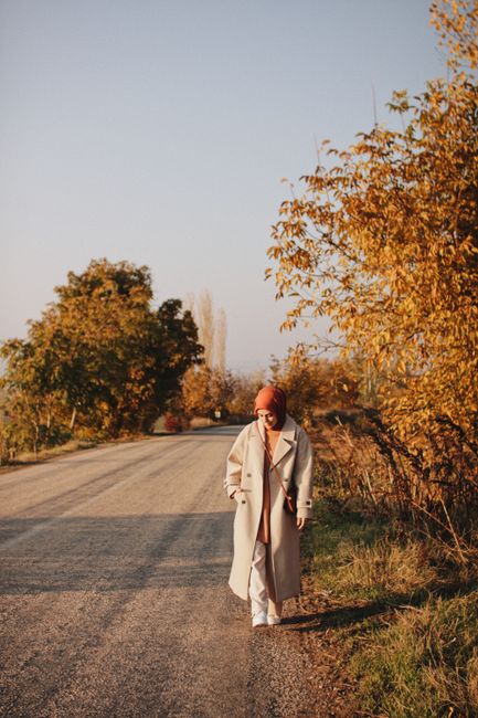 A woman wearing a hijab walks down a quiet country road surrounded by fall foliage and soft light.