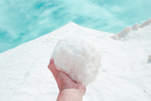 Close-up of a hand holding a snowball against a bright snowy landscape under a clear blue sky.