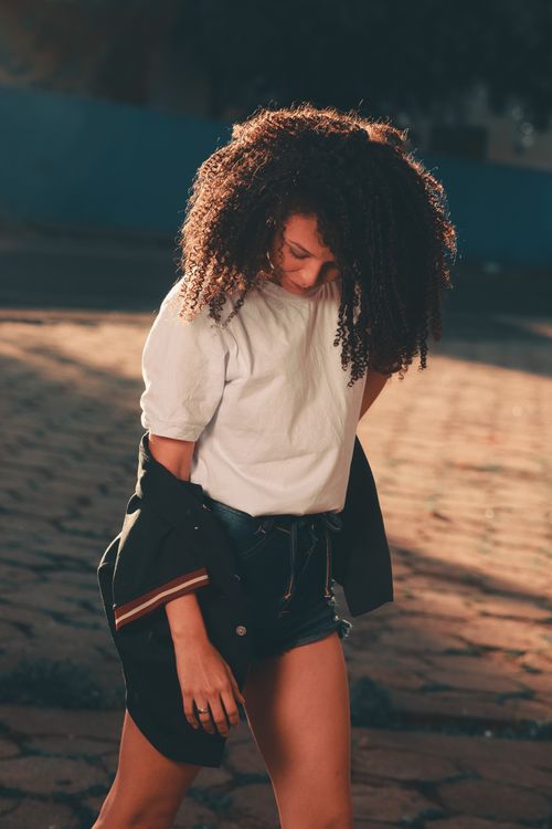 Free A fashionable woman with curly hair in a white shirt poses outdoors in sunlight. Stock Photo