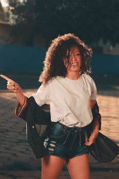 A cheerful woman in denim shorts and a white tee smiling and posing outdoors in warm sunlight.
