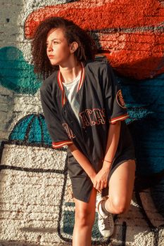 Stylish woman with curly hair posing by a vibrant graffiti wall, filled with sunlight.