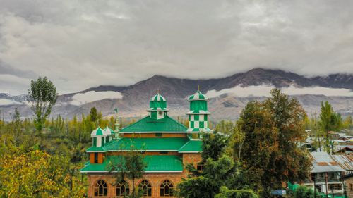 Free A mosque with a green roof surrounded by lush greenery and mountains under a cloudy sky. Stock Photo