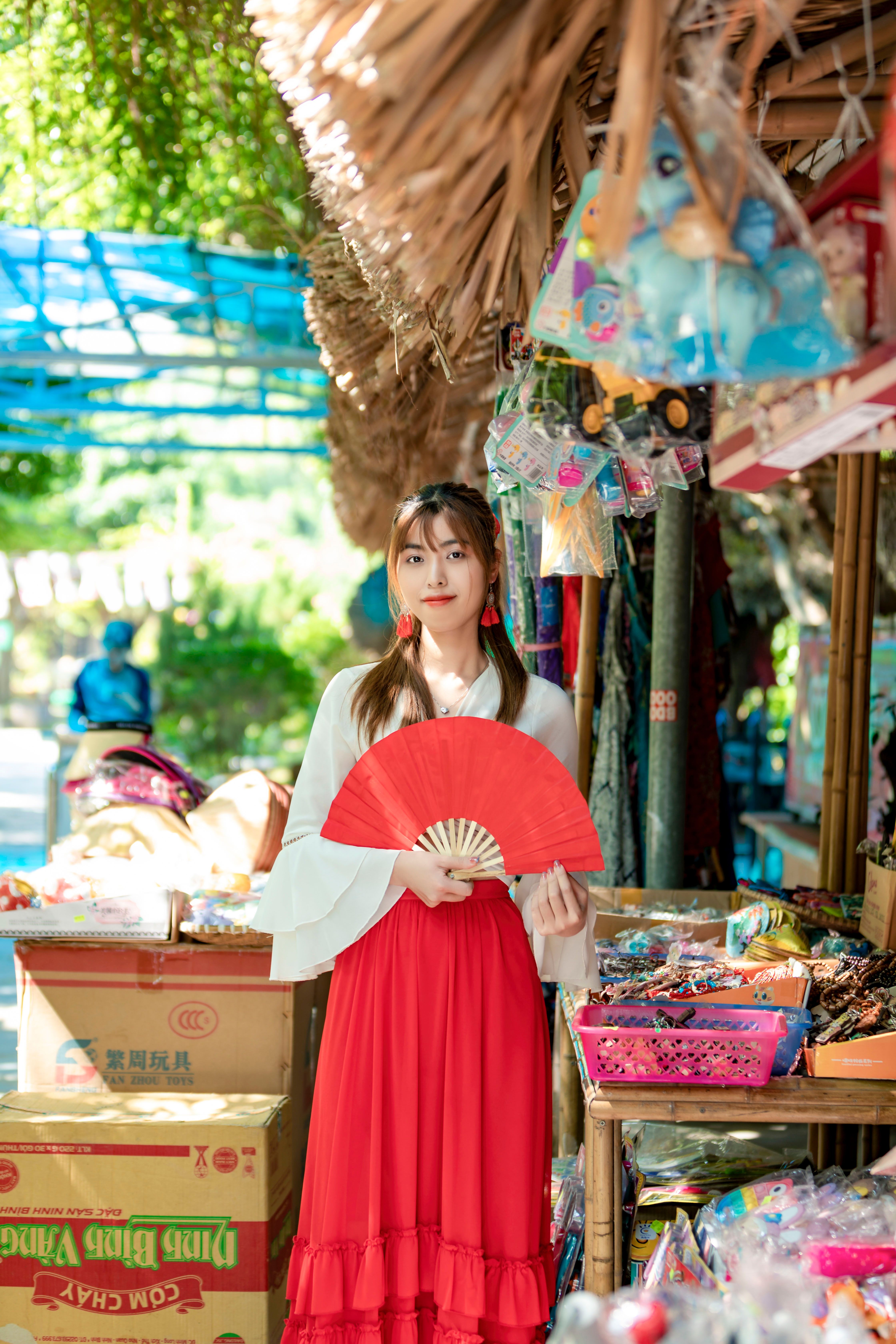 Asian woman in traditional attire holding a red fan at a colorful market stall.