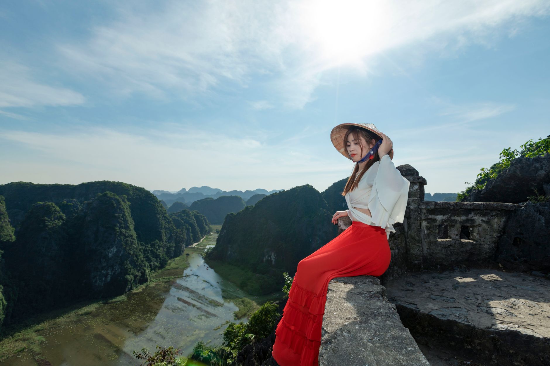 A fashionable woman in a conical hat and red skirt sitting on a stone wall with a stunning valley view.