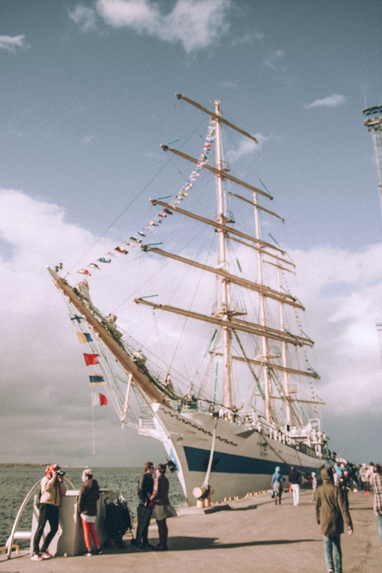 White sailing ship moored in a busy port with crowds of visitors walking along the seafront promenade