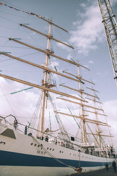 Modern sailing ship with white and blue color with tall masts and people admiring deck of vessel at anchor near harbor in sunny cloudy day