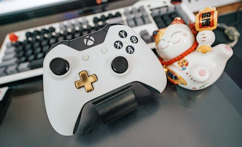 Free White and Black Xbox One Wireless Controller Beside White and Multicolored Ceramic Cat Figurine on Black Surface Stock Photo