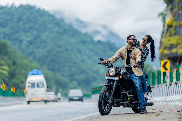 A Couple Sitting On A Motorcycle On The Side Of A Street 