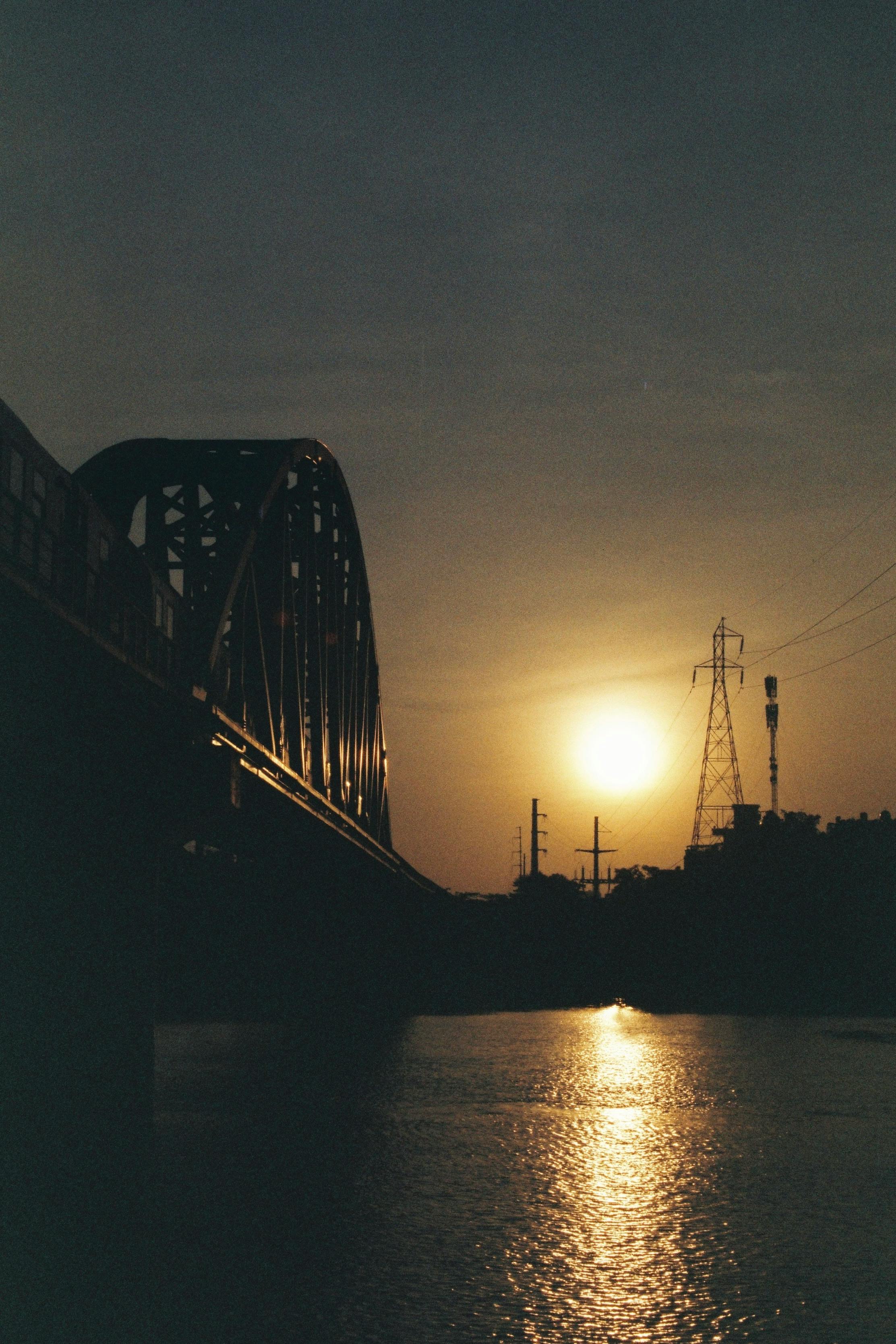 A dramatic silhouette of a bridge spans across a tranquil river at sunset, casting reflections on the water.