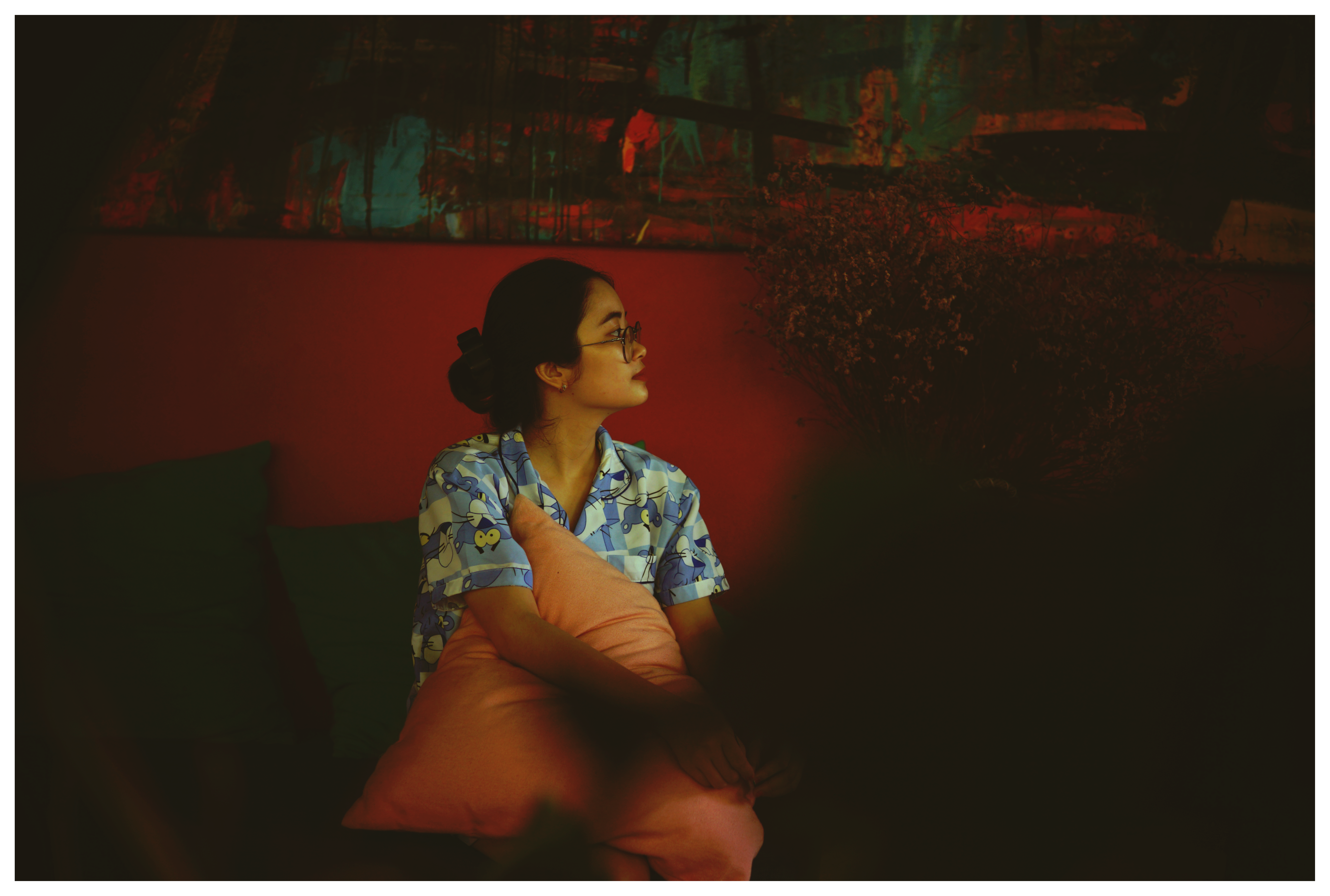 Asian woman with glasses in a dimly lit room, sitting with a pillow.