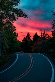 A scenic view of a winding road cutting through forests under a dramatic sunset sky, showcasing natural beauty.