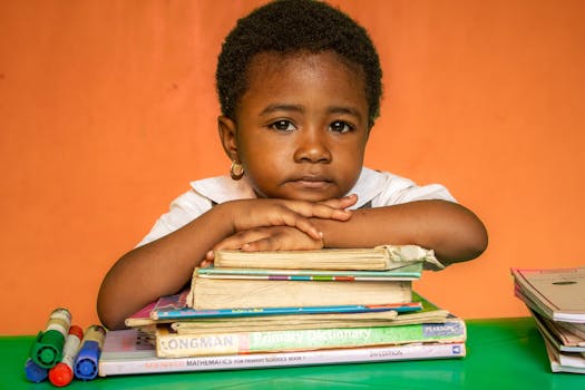Young child resting on a stack of books, surrounded by colorful markers, in a warm classroom setting.