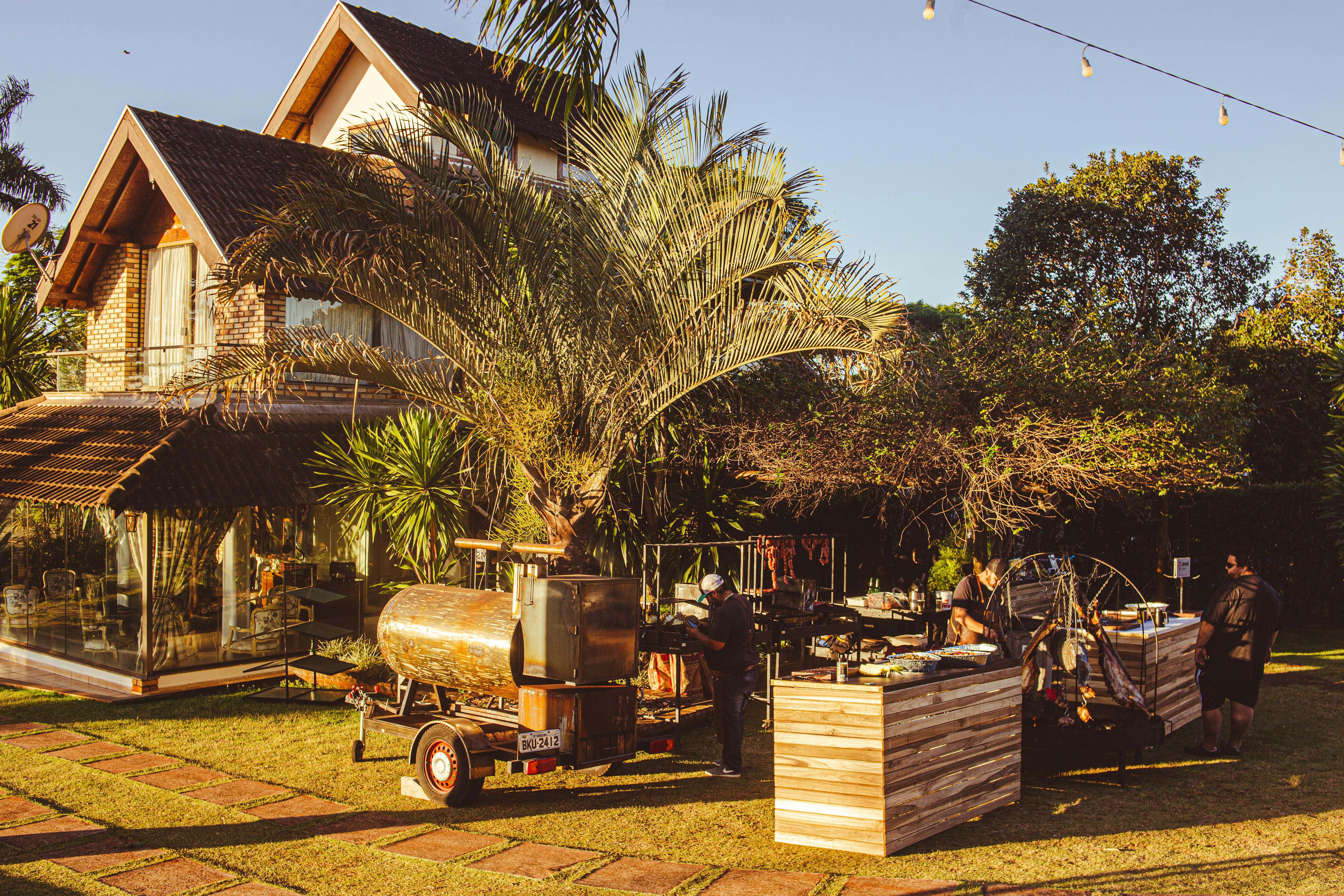 Outdoor BBQ event setup in garden with food truck and tropical trees, during daytime.