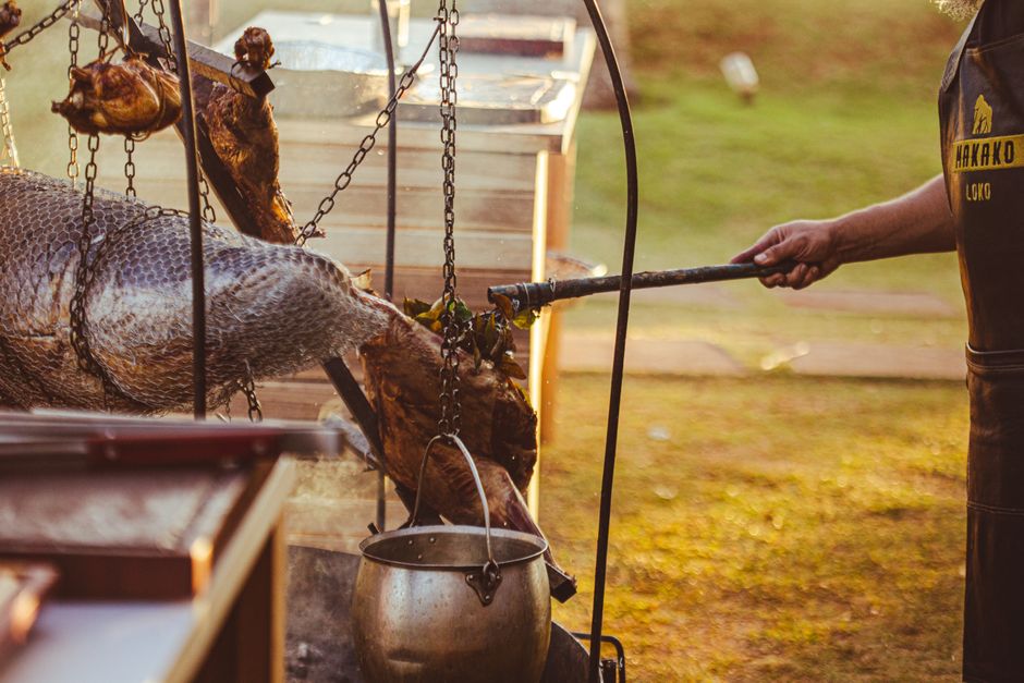 Rustic outdoor cooking scene with grilled meat and fish, including a smoking pot and a man's hand.