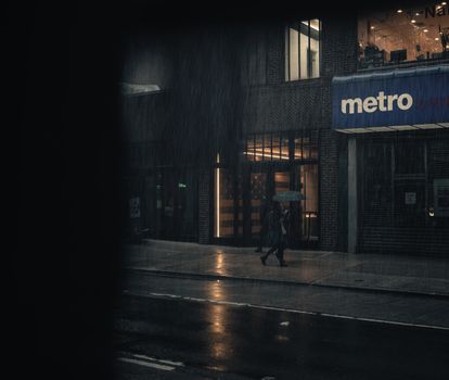 A person walks on a rainy city sidewalk at night, illuminated by streetlights, holding an umbrella.